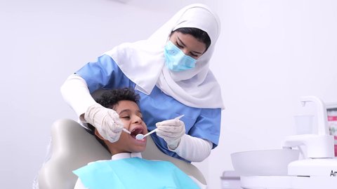 استخدام الأدوات الطبية السنية المعقمة، طبيبه أسنان عربيه خليجيه سعوديه ترتدي اليونيفورم والقفازات والكمامة الطبية تمسك بيدها ادوات طبية، فتي عربي خليجي سعودي في عيادة سنية يتلقى العلاج اللازم، مفهوم العناية بأسنان الأطفال, الأساليب العلاجية المتبعة للعناية بالفم