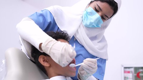 مفهوم العناية بأسنان الأطفال، استخدام الأدوات الطبية السنية المعقمة، طبيبه أسنان عربيه خليجيه سعوديه ترتدي اليونيفورم والقفازات والكمامة الطبية تمسك بيدها ادوات طبية، فتي عربي خليجي سعودي في عيادة سنية يتلقى العلاج اللازم