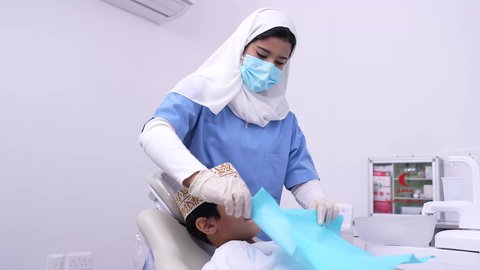 استخدام الأدوات الطبية السنية المعقمة، طبيبه أسنان عربيه خليجيه عمانيه ترتدي اليونيفورم والقفازات والكمامة الطبية تمسك بيدها ادوات طبية، فتى عربي خليجي عماني في عيادة سنية يتلقى العلاج اللازم، مفهوم العناية بأسنان الأطفال, الأساليب العلاجية المتبعة للعناية بالفم