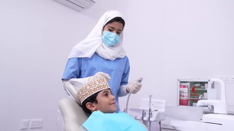 مفهوم العناية بأسنان الأطفال, الأساليب العلاجية المتبعة للعناية بالفم، استخدام الأدوات الطبية السنية المعقمة، طبيبه أسنان عربيه خليجيه عمانيه ترتدي اليونيفورم والقفازات والكمامة الطبية تمسك بيده ادوات طبية، فتى عربي خليجي عماني في عيادة سنية يتلقى العلاج اللازم
