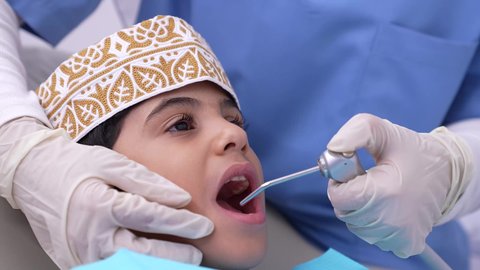 تصوير مقرب لطفل عربي خليجي عماني في عيادة سنية لتقديم الخدمات الصحية, طب وجراحة الفم والأسنان، طبيبه أسنان عربيه خليجيه عمانيه ترتدي القفازات والكمامة الطبية قوم بفحص وتشخيص أسنان طفل مريض، معالجة التسوس وتركيب الحشوات