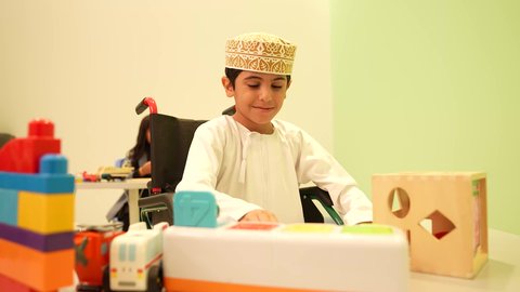 An Omani child wearing a dishdasha and a kuma in kindergartens and nurseries, doing many fun and different activities, a boy with special needs, a group of individual activities for people with disabilities