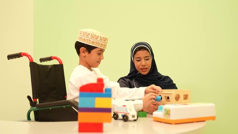 أداء الكثير من الأنشطة الممتعة لذوي الهمم، معلمة عربية عمانيه لرياض أطفال تختص بالاعتناء بطفل ذوي الاحتياجات الخاصة، رياض الأطفال والحاضنات، ولد عربي خليجي عماني مبتسم يرتدي الدشداشه والكمه يقوم بالمهارات والانشطه برفقه معلمته