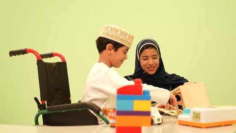 An Omani Arab teacher for a kindergarten specializing in taking care of children with special needs, performing many fun activities for people of determination, kindergartens and nurseries, a smiling Omani Arab Gulf boy wearing a dishdasha and a kuma performing skills and activities with his teacher