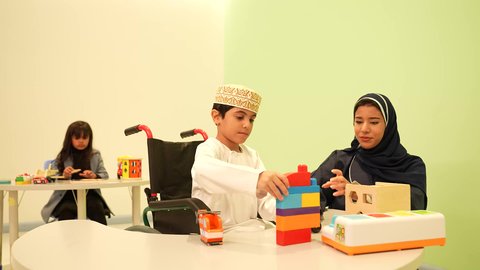 معلمة عربية عمانيه لرياض أطفال تختص بالاعتناء بطفل ذوي الاحتياجات الخاصة، أداء الكثير من الأنشطة الممتعة لذوي الهمم، رياض الأطفال والحاضنات، ولد عربي خليجي عماني مبتسم يرتدي الدشداشه والكمه يقوم بالمهارات والانشطه برفقه معلمته