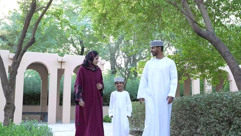 عائلة عمانيه تسير معًا بطمأنينة، ايماءات تعبّر عن المحبة والترابط والسكينه، عائلة تسير معًا في هدوء بين الأشجار، والد عربي خليجي عماني يرتدي الدشداشه والكمه برفقه ابنه الصغير وزوجته، التنزهه في الهواء الطلق