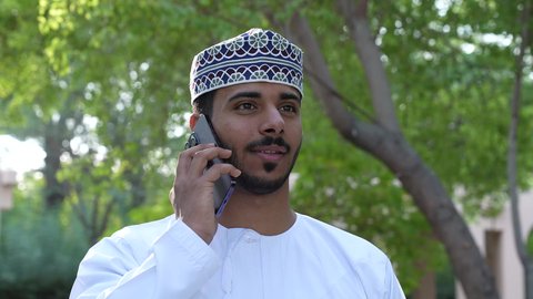 استخدام التقنيات الحديثة، الاستمتاع بالأجواء في إحدى المنتزهات، الاستجمام بالطبيعة واستنشاق الهواء الطلق، الخروج إلى أحد الاماكن العامة للترفيه عن النفس،  رجل عربي خليجي عماني يرتدي الزي العماني يسير فى احدي حدائق سلطنة عمان