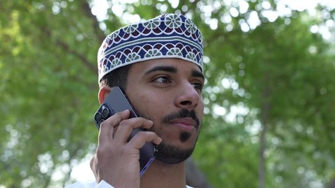 اجراء المكالمات الهاتفية، الاستمتاع بالأجواء في إحدى المنتزهات، الاستجمام بالطبيعة واستنشاق الهواء الطلق، الخروج إلى أحد الاماكن العامة للترفيه عن النفس،  رجل عربي خليجي عماني يرتدي الزي العماني يسير فى احدي حدائق سلطنة عمان