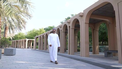 رجل عربي خليجي عماني يرتدي الزي العماني يسير فى احدي حدائق سلطنة عمان ، الخروج إلى أحد الاماكن العامة للترفيه عن النفس،  الاستجمام بالطبيعة واستنشاق الهواء الطلق،الاستمتاع بالأجواء في إحدى المنتزهات
