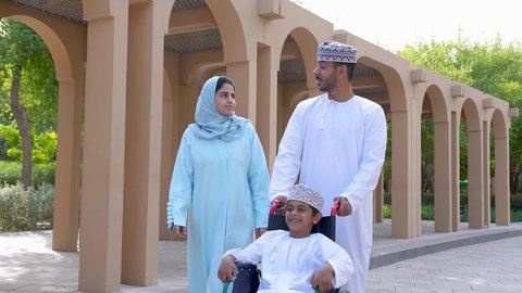 A boy sitting in a wheelchair, an Omani Gulf Arab couple enjoying their son, spending fun time with the family, an outdoor outing, a photo of an Omani Gulf Arab family sitting in a park.