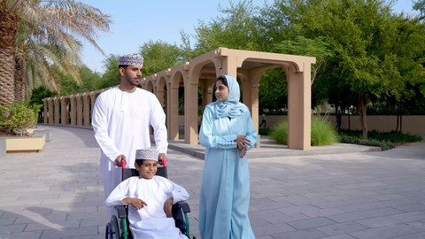 Photograph of an Arab Gulf Omani family sitting in a park, having a picnic and outdoor walk, a boy sitting in a wheelchair, spending fun time with the family, an Arab Gulf Omani couple enjoying themselves with their son.