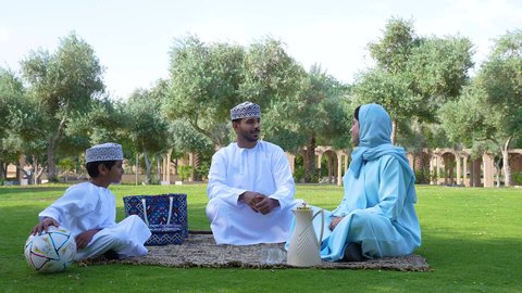 ولد عربي خليجي عماني يرتدي الزي العماني التقليدي يجلس في الحديقة برفقة والديه , ممارسة الأنشطة الترفيهية , اللعب بكرة القدم ، قضاء أوقات متعة مع الأبناء,  إيماءات الفرح والسرور،اجواء الطبيعة الخلابة، خلفية الطبيعة