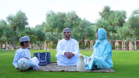ممارسة الأنشطة الترفيهية , اللعب بكرة القدم ، قضاء أوقات متعة مع الأبناء,  إيماءات الفرح والسرور،اجواء الطبيعة الخلابة، ولد عربي خليجي عماني يرتدي الزي العماني التقليدي يجلس في الحديقة برفقة والديه , خلفية الطبيعة