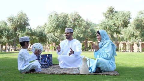 Playing football, having fun with children, gestures of joy and happiness, picturesque natural atmosphere, an Arab Gulf Omani boy wearing traditional Omani clothing sitting in the garden with his parents, practicing recreational activities, nature background