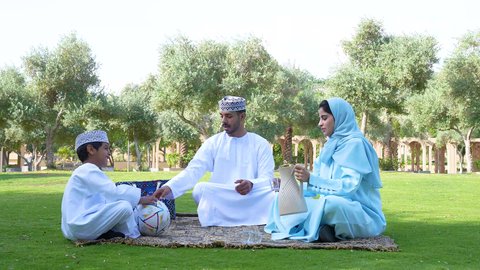 قضاء أوقات متعة مع الأبناء,  إيماءات الفرح والسرور،اجواء الطبيعة الخلابة، ولد عربي خليجي عماني يرتدي الزي العماني التقليدي يجلس في الحديقة برفقة والديه , اللعب بكرة القدم ، ممارسة الأنشطة الترفيهية , خلفية الطبيعة