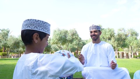 Gestures of joy and happiness, spending fun time with children, picturesque natural surroundings, an Omani Arabian Gulf boy wearing traditional Omani clothing sitting in the park with his father, playing football, engaging in recreational activities, nature background