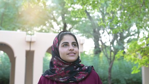 السير في احد الحدائق العامه، امرأه سعوديه ترتدي عباءه وحجاب في وضح النهار وسط جو هادئ ومنعش، سيده عمانيه تقف في مكان طبيعي تحيط به الأشجار الخضراء، التنزهه في الهواء الطلق، ايماءه الاعجاب والفرح