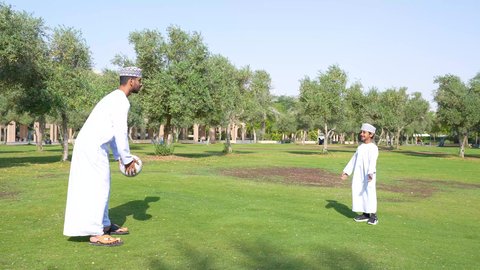 اجواء الطبيعة الخلابة، ولد عربي خليجي عماني يرتدي الزي العماني التقليدي يجلس في الحديقة برفقة والده, اللعب بكرة القدم بإيماءات الفرح والسرور، قضاء أوقات متعة مع الأبناء, ممارسة الأنشطة الترفيهية , خلفية الطبيعة