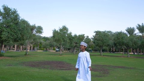 ولد عربي خليجي عماني يرتدي الزي العماني التقليدي يجلس في الحديقة برفقة والده, اللعب بكرة القدم بإيماءات الفرح والسرور، قضاء أوقات متعة مع الأبناء, ممارسة الأنشطة الترفيهية , خلفية الطبيعة