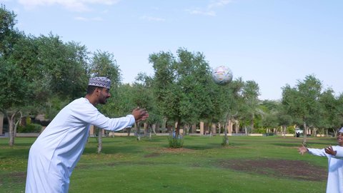 اللعب بكرة القدم بإيماءات الفرح والسرور، قضاء أوقات متعة مع الأبناء, ممارسة الأنشطة الترفيهية ,  ولد عربي خليجي عماني يرتدي الزي العماني التقليدي يجلس في الحديقة برفقة والده, خلفية الطبيعة