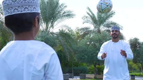 قضاء أوقات متعة مع الأبناء,ممارسة الأنشطة الترفيهية , اللعب بكرة القدم بإيماءات الفرح والسرور، ولد عربي خليجي عماني يرتدي الزي العماني التقليدي يجلس في الحديقة برفقة والده, خلفية الطبيعة