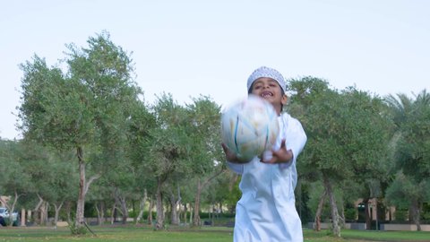 ممارسة الأنشطة الترفيهية وقضاء أوقات متعة مع الأبناء, اللعب بكرة القدم بإيماءات الفرح والسرور، ولد عربي خليجي عماني يرتدي الزي العماني التقليدي يجلس في الحديقة برفقة والده, خلفية الطبيعة