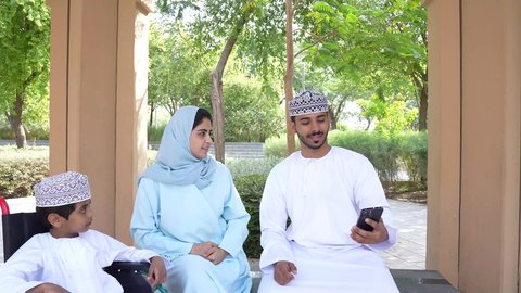 زوجان عربيان خليجيان عمانيان يجلسان فى الحديقة برفقة ولد من ذوى الهمم ، يقومان بإلتقاط صور سيلفي تذكارية بإستخدام الهاتف المحمول، التنزه في حدائق سلطنه عمان، أنشطة عائلية ترفيهية ، الجلوس في الهواء الطلق و الطبيعة الخضراء