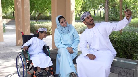 الجلوس في الهواء الطلق و الطبيعة الخضراء ، زوجان عربيان خليجيان عمانيان يجلسان فى الحديقة برفقة ولد من ذوى الهمم ، يقومان بإلتقاط صور سيلفي تذكارية بإستخدام الهاتف المحمول، أنشطة عائلية ترفيهية ، التنزه في حدائق سلطنه عمان