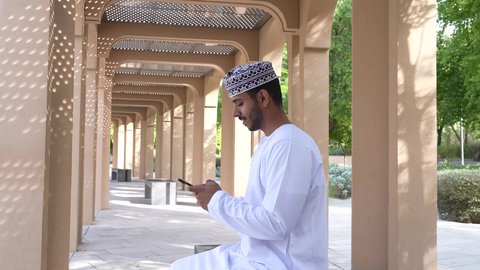 اجواء الطبيعة الخضراء ،رجل عربي خليجي عماني يرتدي الدشداشه والكمه يجلس في الهواء الطلق، قضاء أوقات ممتعة ، استخدام الهاتف المحمول في الرد على اارسائل والمحادثات، التواصل عن بعد، استخدام التقنيات الحديثه