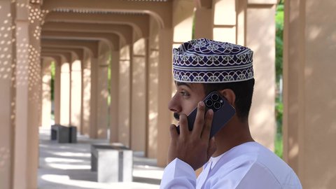 التنزهه في الهواء الطلق ، رجل عربي خليجي عماني يرتدي الدشداشه والكمه يجلس في الحديقة يجري مكالمه هاتفيه، قضاء أوقات ممتعة في الطبيعة الخضراء، استخدام الهاتف المحمول في التصفح عبرالانترنت، التواصل عن بعد، استخدام التقنيات الحديثه
