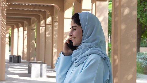 اجراء مكالمه هاتفيه مع الاصدقاء، السير في احد الحدائق العامه، امرأه سعوديه ترتدي عباءه وحجاب في وضح النهار وسط جو هادئ ومنعش، سيده عمانيه تقف في مكان طبيعي تحيط به الأشجار الخضراء، التنزهه في الهواء الطلق، ايماءه الاعجاب والفرح