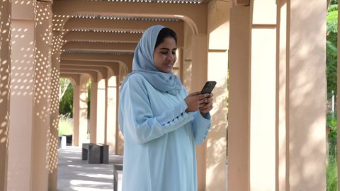 Omani woman sitting in a natural place surrounded by green trees using a mobile phone, Saudi woman wearing an abaya and hijab, calm and refreshing atmosphere, outdoor walk, online communication, use of modern technology