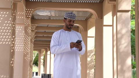 التواصل عن بعد، استخدام التقنيات الحديثه، رجل عربي خليجي عماني يرتدي الدشداشه والكمه يقف في الهواء الطلق، قضاء أوقات ممتعة في الطبيعة الخضراء، استخدام الهاتف المحمول في الرد على الرسائل والمحادثات