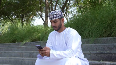 استخدام التقنيات الحديثه، استخدام الهاتف المحمول في الرد على الرسائل والمحادثات، رجل عربي خليجي عماني يرتدي الدشداشه والكمه يجلس في الهواء الطلق، قضاء أوقات ممتعة في الطبيعة الخضراء، ايماءات التفكير والتمعن