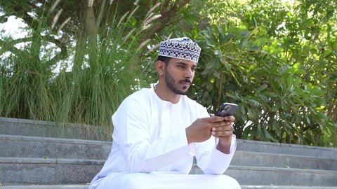 استخدام الهاتف المحمول في الرد على الرسائل والمحادثات، رجل عربي خليجي عماني يرتدي الدشداشه والكمه يجلس في الهواء الطلق، قضاء أوقات ممتعة في الطبيعة الخضراء، استخدام التقنيات الحديثه، ايماءات التفكير والتمعن