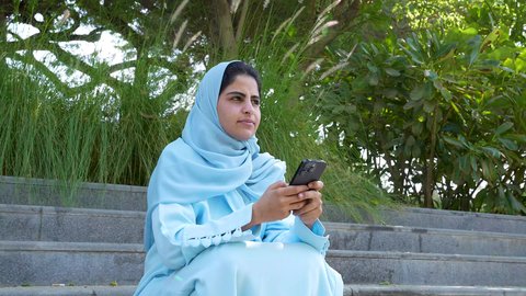النظر بايماءات التأمل في الطبيعه، امراه عمانيه تجلس في مكان طبيعي تحيط به الأشجار الخضراء تستخدم الهاتف، سيده سعوديه ترتدي عباءه وحجاب في وضح النهار ، اجواء هادئه ومنعشه، التنزهه في الهواء الطلق، التواصل عبر الانترنت