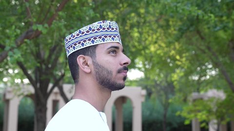 تصوير لرجل عربي خليجي عماني يرتدي الدشداشة والكمة يقف في الحديقة وتحيط به الأشجار الخضراء، النظر إلى الجانب الأيسر والتأمل في الطبيعه، اجواء هادئه ومنعشه