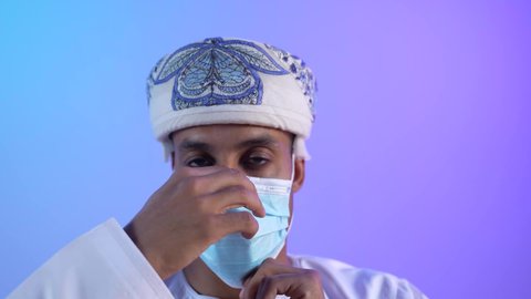اتخاذ التدابير اللازمة للوقاية من فيروس كورونا، النظر الى الكاميرا بايماءات الثقة بالنفس، الاهتمام بالاناقة والمظهر الخارجي، اتباع الإجراءات الاحترازية لمنع انتشار العدوى، بورتريه مقرب لشاب عربي خليجي عماني يرتدي الدشداشة والكمه يضع الكمامة الطبية لتجنب الاصابة بالامراض، خلفية زرقاء