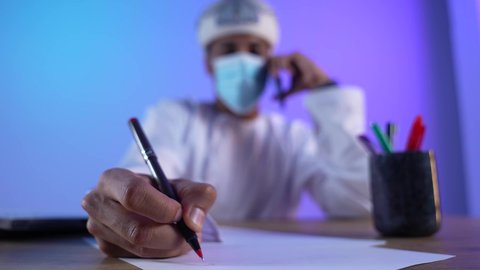 مفهوم ادارة الاعمال عن بعد، مهن ووظائف مكتبيه عمانية, استخدام الأجهزة الحديثة والتقنيات المتطورة، بورتريه مقرب لرجل عربي خليجي عماني يرتدي الدشداشة والكمه العمانية التقليدية يقوم باجراء مكالمة هاتفية عبر الهاتف المحمول و كتابة شيء ما, ارتداء الكمامة لإجراءات الوقاية من فايروس كورونا، خلفية زرقاء