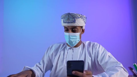 مفهوم ادارة الاعمال عن بعد، مهن ووظائف مكتبيه عمانية, استخدام الأجهزة الحديثة والتقنيات المتطور، بورتريه مقرب لرجل عربي خليجي عماني يرتدي الدشداشة و الكمة يقوم بتصفح الهاتف المحمول، ارتداء الكمامة لإجراءات الوقاية من فايروس كورونا، خلفية زرقاء