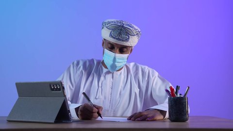 دوين ملاحظات ، التسويق الإلكتروني ، مفهوم التكنولوجيا وإستخدام الأجهزة التقنية الحديثة ، العمل عن بعد ، مهن و وظائف ، الكتابة والعمل على شاشة الجهاز التقني، بورتريه مقرب لرجل عربي خليجي عماني يرتدي الدشداشة و الكمة يقوم بكتابة شيء ما، ارتداء الكمامة لإجراءات الوقاية من فايروس كورونا، خلفية زرقاء