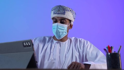 متابعة الأفلام والمسلسلات الممتعة، استخدام التكنولوجيا و التقنيات الحديثة, تصفح مواقع التواصل الاجتماعي، المحادثة مع الأهل والأصدقاء، بورتريه مقرب لرجل عربي خليجي عماني يرتدي الدشداشة و الكمة يحمل بيده جهاز التابلت، ارتداء الكمامة لإجراءات الوقاية من فايروس كورونا، خلفية زرقاء