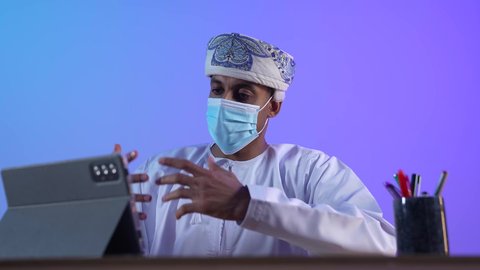 المحادثة مع الأهل والأصدقاء، استخدام التكنولوجيا و التقنيات الحديثة, تصفح مواقع التواصل الاجتماعي، متابعة الأفلام والمسلسلات الممتعة، بورتريه مقرب لرجل عربي خليجي عماني يرتدي الدشداشة و الكمة يحمل بيده جهاز التابلت، ارتداء الكمامة لإجراءات الوقاية من فايروس كورونا، خلفية زرقاء