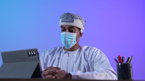 استخدام التكنولوجيا و التقنيات الحديثة, تصفح مواقع التواصل الاجتماعي، متابعة الأفلام والمسلسلات الممتعة، المحادثة مع الأهل والأصدقاء، بورتريه مقرب لرجل عربي خليجي عماني يرتدي الدشداشة و الكمة يحمل بيده جهاز التابلت، ارتداء الكمامة لإجراءات الوقاية من فايروس كورونا، خلفية زرقاء