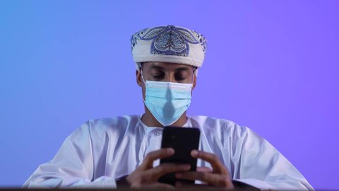 استخدام الأجهزة الحديثة والتقنيات المتطورة،مفهوم ادارة الاعمال عن بعد، مهن ووظائف مكتبيه عمانية، بورتريه مقرب لرجل عربي خليجي عماني يرتدي الدشداشة والكمه العمانية يجلس على المكتب و يقوم بتصفح الهاتف المحمول، ارتداء الكمامة لإجراءات الوقاية من فايروس كورونا، خلفية زرقاء