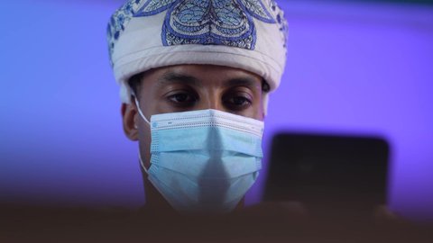 مفهوم ادارة الاعمال عن بعد، مهن ووظائف مكتبيه عمانية, استخدام الأجهزة الحديثة والتقنيات المتطورة، بورتريه مقرب لرجل عربي خليجي عماني يرتدي الدشداشة والكمه العمانية يجلس على المكتب و يقوم بتصفح الهاتف المحمول، ارتداء الكمامة لإجراءات الوقاية من فايروس كورونا، خلفية زرقاء