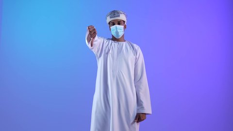 الشعور بالإنزعاج من شيء ما، مفهوم الرفض والفشل، بورتريه لشاب عربي خليجي عماني يرتدي الدشداشة والكمه يخفض ابهامه بايماءات الرفض وعدم القبول، تعابير التوتر وعد الرضا،رتداء الكمامة لإجراءات الوقاية من فايروس كورونا، خلفية زرقاء