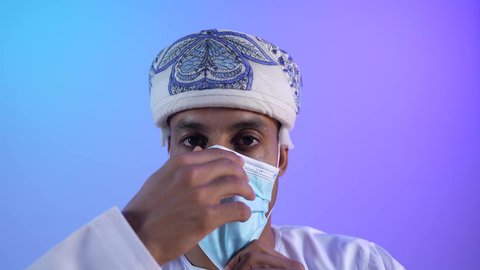 النظر الى الكاميرا بايماءات الثقة بالنفس، الاهتمام بالاناقة والمظهر الخارجي، اتباع الإجراءات الاحترازية لمنع انتشار العدوى، بورتريه مقرب لشاب عربي خليجي عماني يرتدي الدشداشة والكمه يضع الكمامة الطبية لتجنب الاصابة بالامراض، اتخاذ التدابير اللازمة للوقاية من فيروس كورونا، خلفية زرقاء