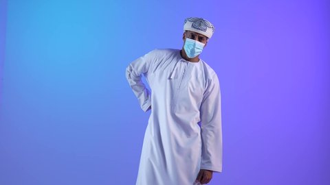 الأوجاع والآلام العصبية، طلب المساعده والحاجه للعلاج، التهاب العضلات والمفاصل، بورتريه لشاب عربي خليجي عماني يرتدي الدشداشة و الكمه يضع يده على ظهره بايماءات الألم والتعب والأرهاق، حالة اسعافية طارئة،ارتداء الكمامة لإجراءات الوقاية من فايروس كورونا، خلفية زرقاء