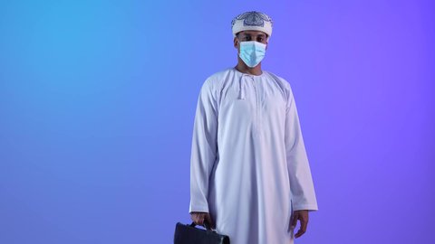 الاهتمام بالاناقة والمظهر الخارجي، بورتريه لرجل أعمال عربي خليجي عماني يرتدي الدشداشة والكمة والكمامة الواقية يحمل حقيبة العمل، الالتزام باجراءات الوقاية من فايروس كورونا، خلفية زرقاء
